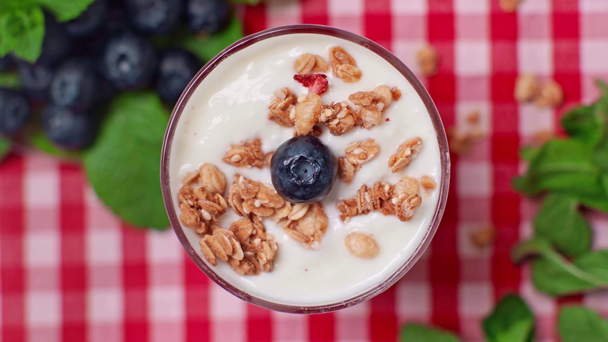 bovenaanzicht van de mens die bosbessen bovenop yoghurt zet met granola in glas - Video