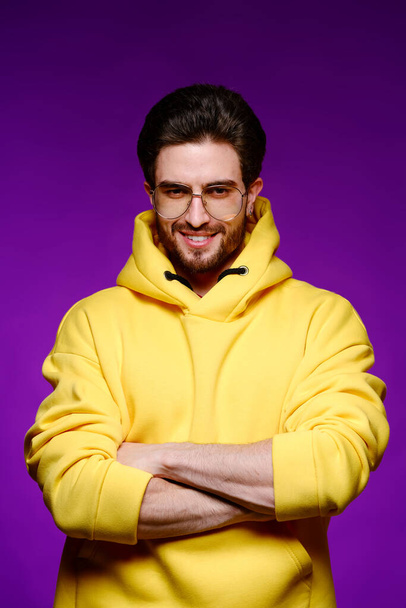 A young man of 25-30 years in glasses and a yellow sweatshirt emotionally poses on a purple background.  - Foto, Bild
