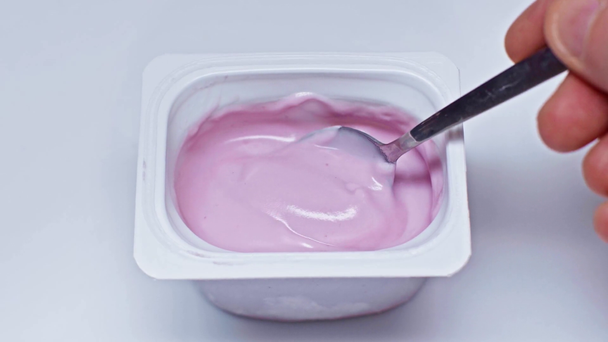 przycięty widok człowieka trzymającego łyżkę w pobliżu pojemnika z jogurtem na białym - Materiał filmowy, wideo