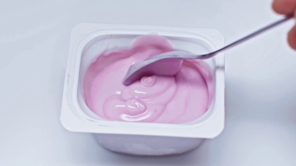 Powolny ruch człowieka trzymającego łyżkę w pobliżu pojemnika z jogurtem na białym - Materiał filmowy, wideo