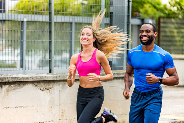 Jeune couple se souriant tout en faisant du jogging dans la rue, dehors pour courir. Afro américain homme et belle fille blonde en vêtements de sport courir dans la rue de la ville ensemble - Santé et remise en forme - Photo, image