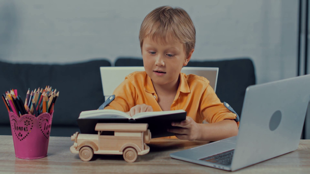 roztomilý chlapec čtení knihy v blízkosti notebooku, zatímco e-learning doma - Záběry, video