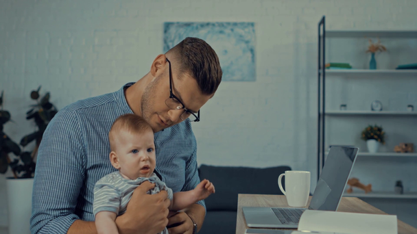 handsome freelancer holding in arms cute infant son and using laptop  - Footage, Video