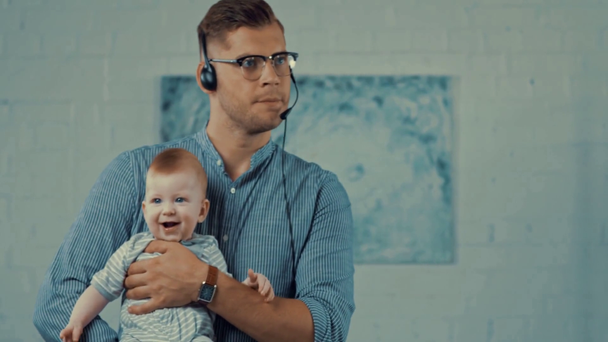 在宅勤務中に腕の幼児の息子を抱えるハンサムなオペレーター - 映像、動画