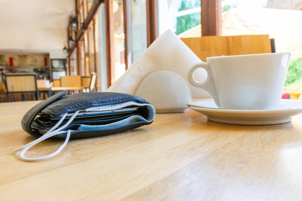 Zwarte portemonnee met gezichtsmasker binnenin met witte beker, servetten in een houder rond in cafe zonder mensen. Veiligheidsregels en -voorschriften in de wereld na de corona pandemie. Economie in 2020 - Foto, afbeelding