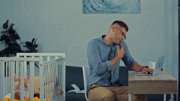 pai usando laptop e falando no smartphone perto berço do bebê com o bebê
 - Filmagem, Vídeo