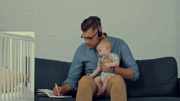 όμορφος χειριστής που κρατά το παιδί του ενώ εργάζεται από το σπίτι - Πλάνα, βίντεο