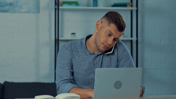 guapo freelancer hablando en el teléfono inteligente cerca del ordenador portátil en casa
 - Metraje, vídeo