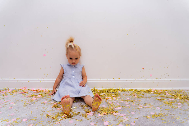 Cute caucasuian blond baby girl sits on a rug with a lot of gold tinsels after celebration holiday, bare feet. - Foto, immagini