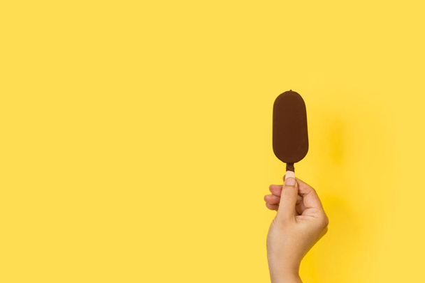 Woman hand holding a dark chocolate dipped ice lolly on a yellow background - Fotó, kép
