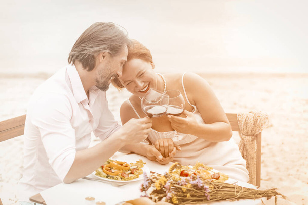 Charming Mature couple drinking wine at sea side. Two smiling Caucasian people spend time together in beach cafe. Love concept. Anniversary concept. Tinted image - 写真・画像