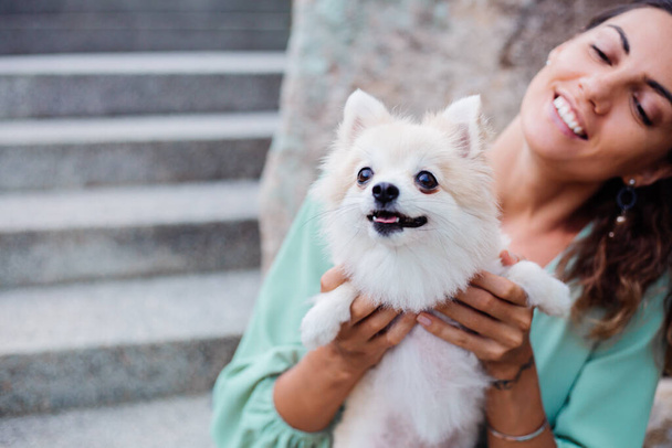 A kültéri portré göndör európai cserzett nő tartja boldog kisállat kutya pomerániai köpködés. Ezüst karkötő. Bolyhos fehér kiskutyával töltöm az időmet. Boldog pillanatok háziállatokkal. Egzotikus háttér. - Fotó, kép