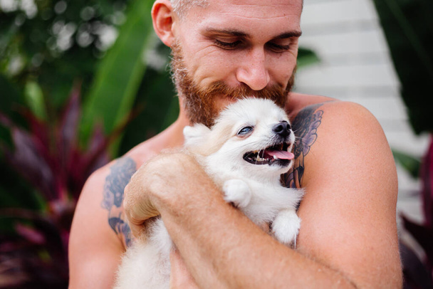 Young handsome bearded brutal tattooed happy man holds pomeranian spitz, playing with lovely pet. Male spending time outside in park with cute fluffy dog puppy. Tropical exotic background. - Fotoğraf, Görsel