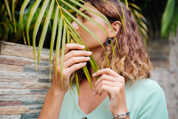 Cerrar retrato exterior de mujer europea bronceada con pelo corto en pendientes redondos de joyería con fondo tropical de hoja de palma
. - Foto, imagen