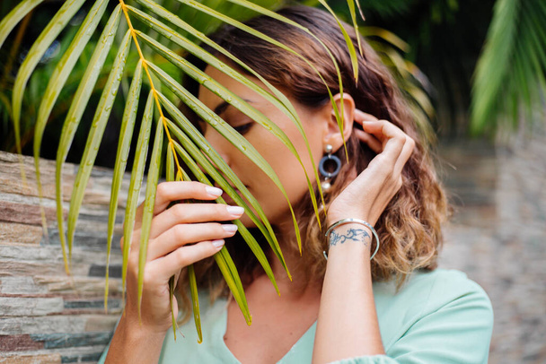 Fechar retrato ao ar livre de mulher europeia bronzeada com cabelo curto em brincos redondos de jóias com folha de palma fundo tropical
. - Foto, Imagem