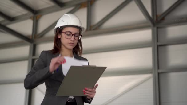 Nuori nainen kypärässä dokumenttien kanssa rakennustyömaalla. Pukumies pitää kirjaa arkkitehtuurista. Tyttö katselee ympärilleen.. - Materiaali, video