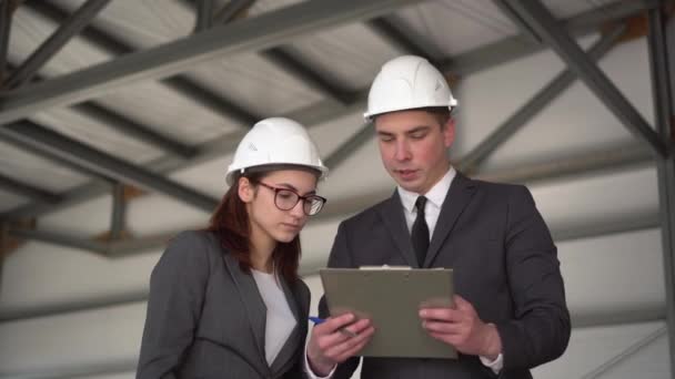 Young man and woman in helmets with documents at a construction site. Businessmen in suits conclude an agreement. A woman signed documents. - Footage, Video