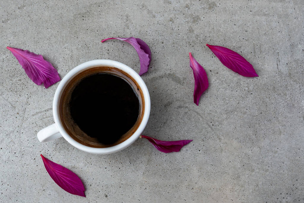 Una taza de café de cerámica blanca sobre una mesa de hormigón gris con pétalos de flores de color rojo violeta. Vista superior
. - Foto, Imagen