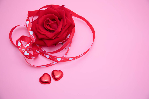 Detailní záběr krásné červené růže s červenou a bílou srdíčkovou stuhou na růžovém pozadí se dvěma červenými sladkými srdíčky. Valentýn, výročí, svatební koncept. - Fotografie, Obrázek