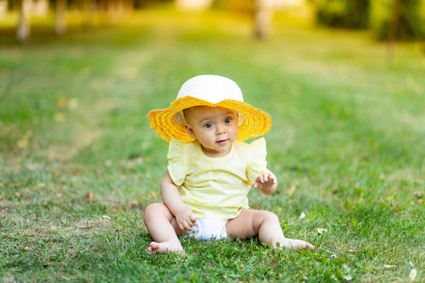 Девочка 8 месяцев сидит летом на зеленой траве в желтом летнем платье и шляпе, место для текста. - Фото, изображение