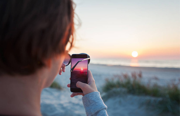 Chica tomando disparar a espectacular puesta de sol de fondo en la playa con smartphone y gafas de sol. Imagen horizontal multicolor vibrante al aire libre con filtro
 - Foto, imagen