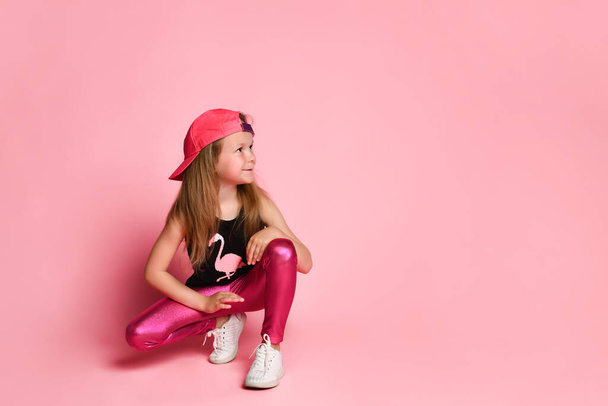 Charming active preschool girl in a trendy fashionable outfit, playful and smiling, squatting and looking to the side on a pink background - Photo, Image