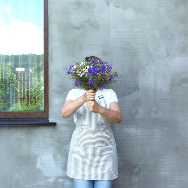 Önlüklü kız yüzünü bir buket çayır çiçeğinin arkasına saklar. Pencere gökyüzünü ve çiti yansıtıyor. F - Fotoğraf, Görsel