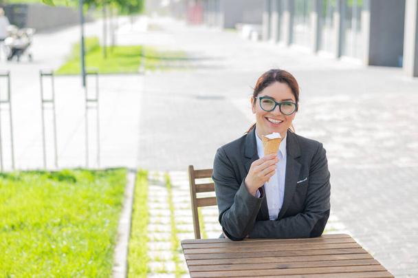Smiling woman enjoys eating an ice cream cone while sitting at a wooden table outdoors. Portrait of a girl in a business suit for a break in a street cafe on a hot summer day. - Foto, Imagen