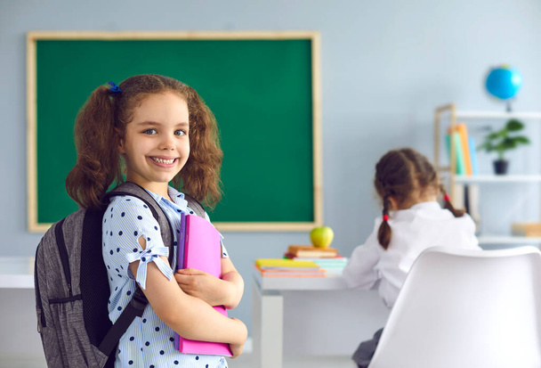 Back to school. Smiling schoolgirl with a book in her hand looks at the camera while standing in class. - Foto, imagen