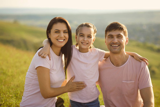 Portrait of positive young family looking at camera in countryside. Cute girl hugging her parents on summer vacation - Фото, зображення