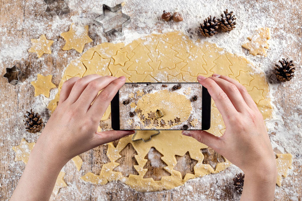 Top view of hands of a person making photos with the mobile phone to the raw dough to make Christmas tree shaped cookies and star shapes. On a floured table with pineapples. Christmas desserts - 写真・画像