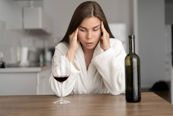 Θλιβερή κουρασμένη γυναίκα πίνει κρασί στο σπίτι, μόνη, κάθεται σε ένα τραπέζι στην κουζίνα, νιώθει κατάθλιψη. Μοναξιά και άγχος. Στρες κρίσης - Φωτογραφία, εικόνα