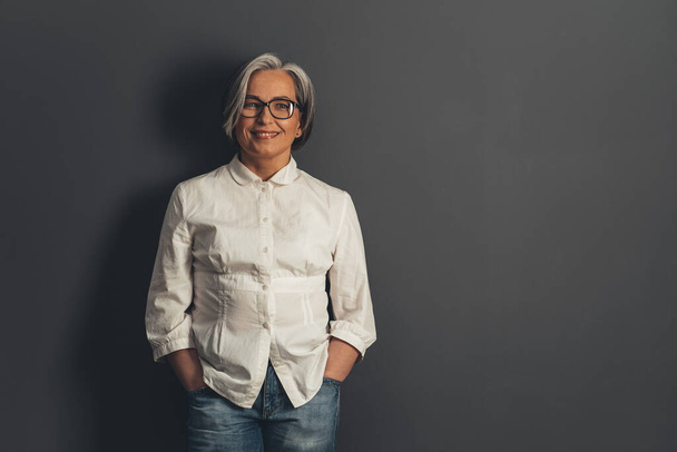 Confident mature woman toothy smiling while standing hands in pockets. Happy senior woman cut out on gray background with copy space at right side. Tinted image - 写真・画像