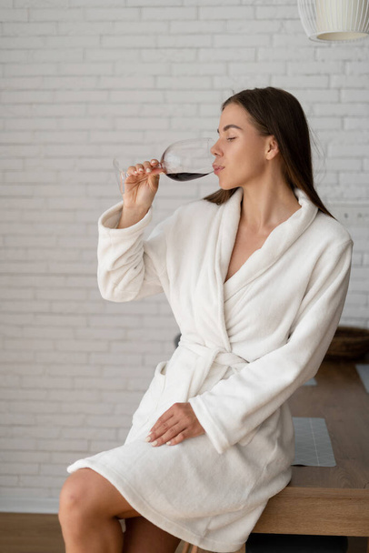 Belle n femme en manteau blanc profitant d'un verre de vin rouge sec assis sur la table de la cuisine de l'hôtel. - Photo, image