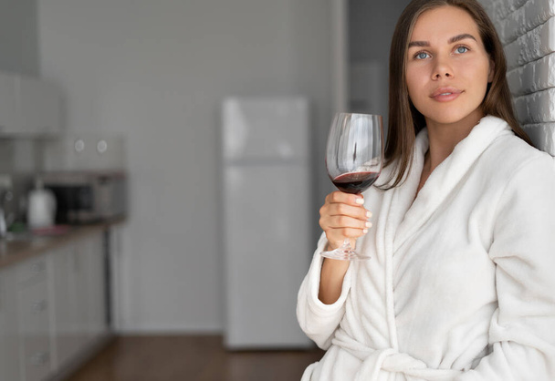 fiatal gyönyörű nő egy pohár borral egy hotelszobában. Egyedül pihen. Igyál bort és nézz ki az ablakon. Fehér fürdőköpeny. - Fotó, kép