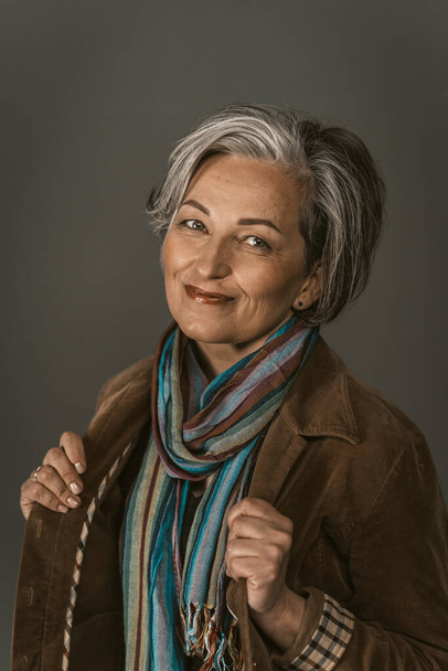 Smiling mature woman in casual. Female portrait on gray. Lady wearing colorfull scarf and jacket smiles looking at camera. Tinted image - Φωτογραφία, εικόνα
