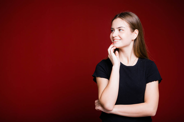 Beautiful American woman in a black T-shirt holds a hand near her face and looks away smiling on a red background - Photo, image