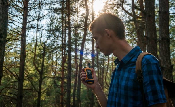 A young man in a blue plaid shirt with a walkie-talkie in his hand stands in a coniferous forest. - Foto, Imagem