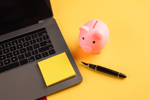Moneybox near laptop and yellow sticky note, place for text. Finance and budget concept. Piggy bank in pink color with gadgets and stationery on colorful background - Foto, imagen