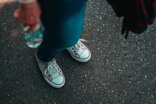 Jeune femme portant des baskets blanches et un jean tenant une bouteille en plastique avec espace de copie. Millénaire vêtu occasionnellement avec des chaussures à la mode debout sur l'asphalte gris dans la ville - Photo, image