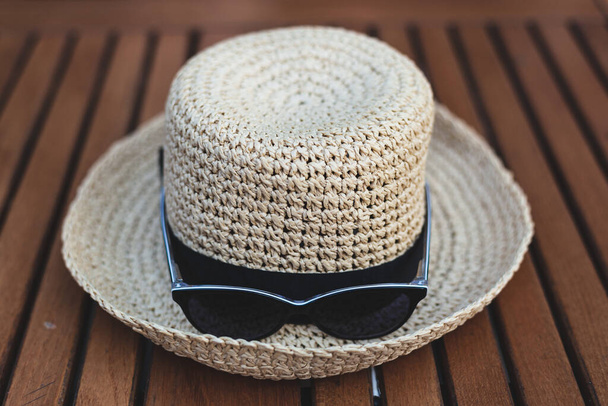 Žlutý slaměný klobouk se stuhou a černými slunečními brýlemi umístěnými na dřevěném stole venku. Základní letní doplňky pro ochranu proti teplu a jasnému slunečnímu svitu - Fotografie, Obrázek