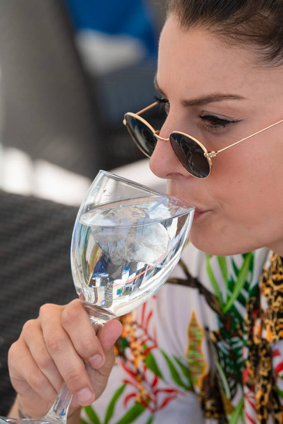 Zbliżenie pięknej kobiety w okularach pijącej ze szklanki wody z kostkami lodu na nieostrym tle. Koncepcja zdrowego stylu życia i wakacji. - Zdjęcie, obraz