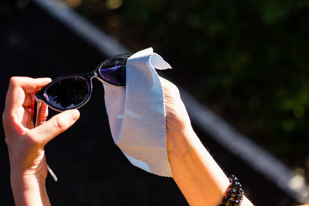 Osoba čištění černé sluneční brýle s hladkou modrou látkou venku. Mladá žena drží letní brýle a zároveň odstraňuje prach z nich měkkou tkaninou - Fotografie, Obrázek