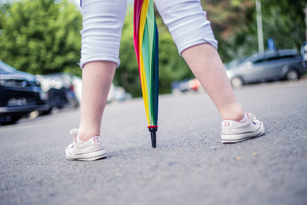 Egy fiatal nő fehér rövidnadrágban és tornacipőben, színes esernyőt tartva a lábai között. Személy láb séta a város utcáján szivárványszínű napernyővel - Fotó, kép