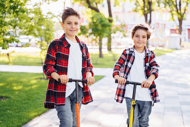 Due attraenti fratelli ragazzi europei, indossando camicie di flannek a scacchi rossi e bianchi, in piedi su scooter nel parco. Ridono, sorridono, si abbracciano e si divertono. Mostrando grandi dita su segno. - Foto, immagini