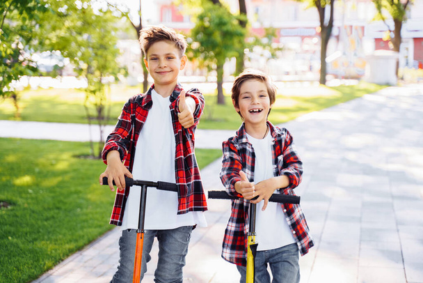Two attractive european boys brothers, wearing red and white checkered flannek shirts, standing on scooters in the park. They laughing, smiling, hugging and having fun. Showing big fingers up sign. - Фото, изображение