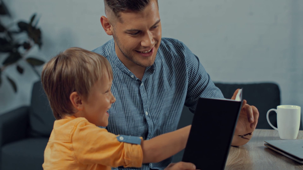 cheerful father showing notebook to cute son at home - Imágenes, Vídeo