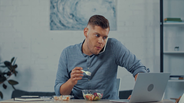 απασχολημένος freelancer τρώει σαλάτα και μιλώντας στο smartphone, ενώ χρησιμοποιώντας φορητό υπολογιστή - Πλάνα, βίντεο