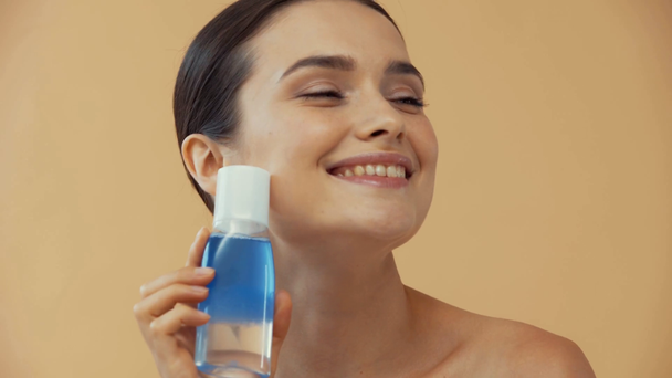 Nudo donna sorridente in possesso di bottiglia con struccante isolato su beige - Filmati, video