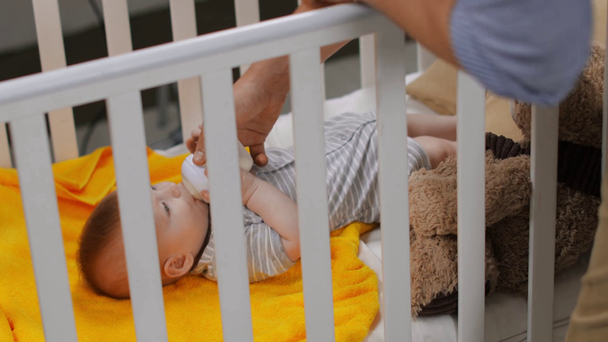 father holding baby bottle and feeding infant son in crib - Video